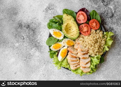 Delicious healthy lunch consisting of chicken, avocado, quinoa, eggs, tomatoes, lettuce and greens on a black plate on a concrete background