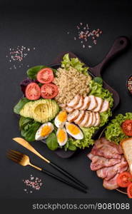 Delicious healthy lunch consisting of chicken, avocado, quinoa, eggs, tomatoes, lettuce and greens on a black plate on a concrete background