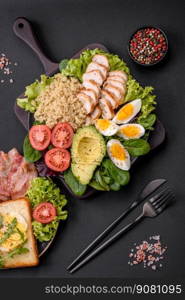 Delicious healthy lunch consisting of chicken, avocado, quinoa, eggs, tomatoes, lettuce and greens on a black plate on a concrete background