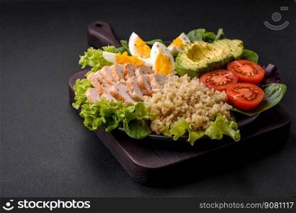 Delicious healthy lunch consisting of chicken, avocado, quinoa, eggs, tomatoes, lettuce and greens on a black plate on a concrete background