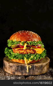 Delicious homemade grilled burger with turkey cutlet with cheese, cheese sauce, tomato, cucumber, salad on wooden board, on dark background. Close up, selective focus with copy space, vertical frame