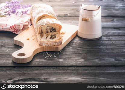 Delicious homemade sponge cake with poppy seed and walnuts, sliced on a trencher, near an antique bucket with milk, on a black wooden table, in the morning light.