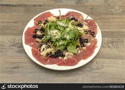 Delicious Italian recipe for carpaccio with parmesan cheese, slices of black olives, olive oil, arugula and basil, beef and capers in vinegar