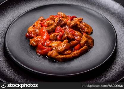
Delicious juicy meat with hot peppers and sauce on a black ceramic plate on a dark concrete background