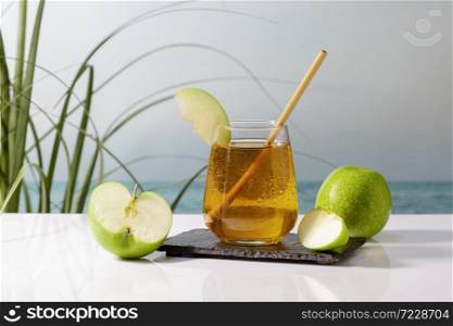 Delicious looking glass of apple juice with a straw and an apple slice on a dark plate surrounded by apples on an out of focus background. Vegan and healthy food concept.. Delicious glass of apple juice with a straw and an apple slice on a dark plate.healthy food concept