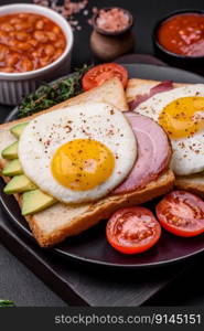 Delicious nutritious English breakfast with fried eggs, tomatoes and avocado with spices and herbs on a dark concrete background