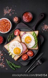 Delicious nutritious English breakfast with fried eggs, tomatoes and avocado with spices and herbs on a dark concrete background
