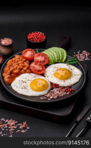 Delicious nutritious English breakfast with fried eggs, tomatoes and avocado with spices and herbs on a dark concrete background