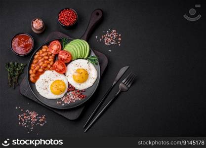 Delicious nutritious English breakfast with fried eggs, tomatoes and avocado with spices and herbs on a dark concrete background
