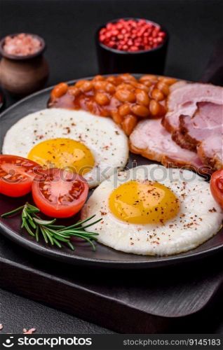 Delicious nutritious English breakfast with fried eggs, tomatoes, bacon and beans on a dark concrete background