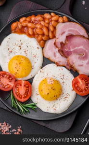 Delicious nutritious English breakfast with fried eggs, tomatoes, bacon and beans on a dark concrete background