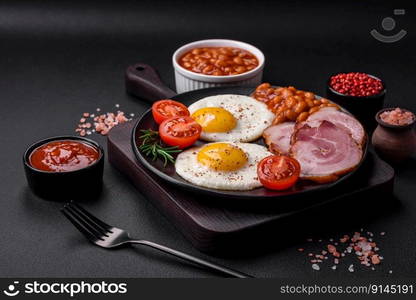 Delicious nutritious English breakfast with fried eggs, tomatoes, bacon and beans on a dark concrete background