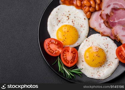 Delicious nutritious English breakfast with fried eggs, tomatoes, bacon and beans on a dark concrete background