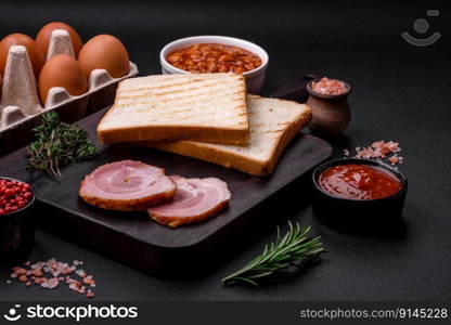 Delicious nutritious English breakfast with fried eggs, tomatoes, bacon and beans on a dark concrete background