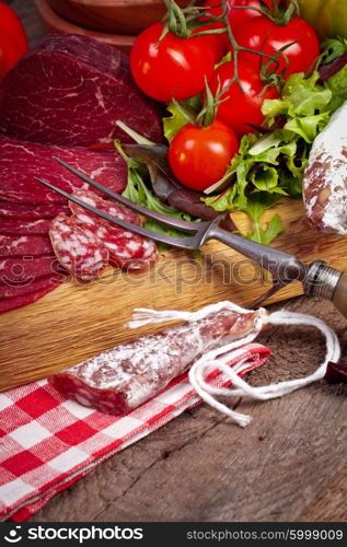 Delicious prosciutto and salami slices on wooden chopping board with vegetables and herbs. Culinary traditional ham.