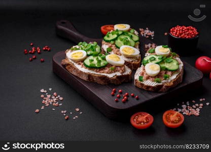 Delicious sandwiches consisting of grilled toast, canned tuna, cream cheese, cucumber and boiled quail eggs on a dark concrete background