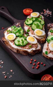 Delicious sandwiches consisting of grilled toast, canned tuna, cream cheese, cucumber and boiled quail eggs on a dark concrete background