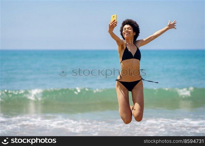 Delighted African American female tourist in swimwear jumping with raised arm while taking self portrait on smartphone near sea in tropical resort. Happy black woman taking selfie while jumping on coast