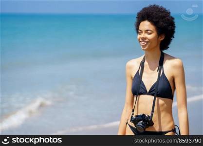 Delighted African American female traveler in bikini with professional photo camera standing near rippling sea on sunny day and looking away. Smiling black woman with photo camera near sea