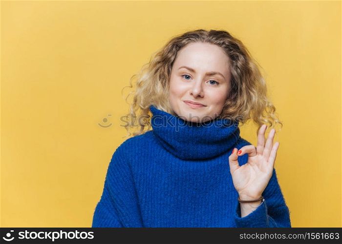 Delighted attractive young female with blonde curly hair, satisfied expression, shows ok sign, demonstrates approval concerning something, isolated over yellow background. That`s fine! All right