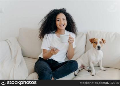 Delighted dark skinned woman poses in modern apartment, sits on comfortable sofa with pet, drinks coffee, uses mobile phone for online communication, being in good mood, flipping news, uses app