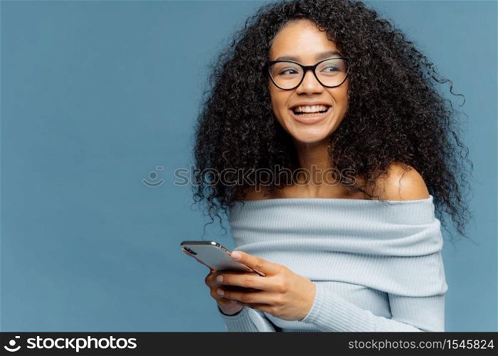 Delighted dark skinned woman with crisp hair, looks away, wears spectacles, uses modern cell phone for chating online, dressed in casual jumper, isolated over blue background with copy space