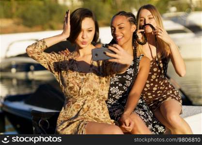 Delighted diverse female friends in dresses taking self portrait on smartphone while sitting together in wharf near river with boats. Positive multiracial women taking selfie in port