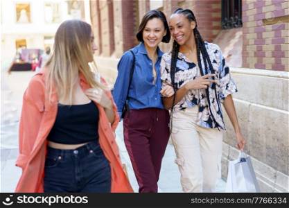 Delighted diverse female friends with shopping bags looking at each other while walking together on street after shopping in city. Joyful multiracial women with shopping bags in city