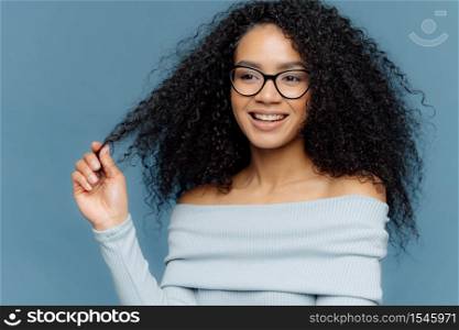 Delighted female holds curl, has bushy hair, being in high spirit, enjoys free time, wears fashionable light blue sweater, shows bare shoulders, stands indoor. People, beauty and happiness concept