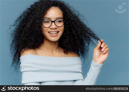 Delighted female holds curl, has bushy hair, being in high spirit, enjoys free time, wears fashionable light blue sweater, shows bare shoulders, stands indoor. People, beauty and happiness concept
