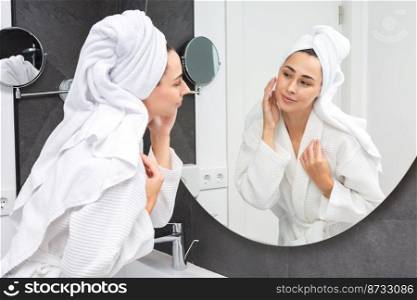 Delighted female in bathrobe applying moisturizing cream on face while standing against mirror in bathroom and looking at mirror. Content woman applying cream in light bathroom