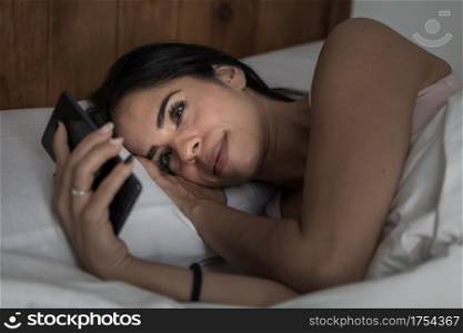 Delighted female in pajama lying under blanket in bed and surfing Internet via mobile phone before sleep. Content woman lying in bed and browsing smartphone