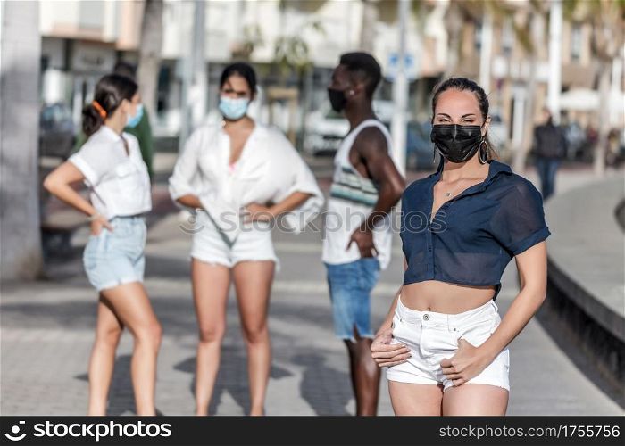 Delighted female in protective mask and summer wear standing in city on background of group of diverse friends while spending time together on Lanzarote. Content woman standing on street against company of friends