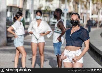 Delighted female in protective mask and summer wear standing in city on background of group of diverse friends while spending time together on Lanzarote. Content woman standing on street against company of friends