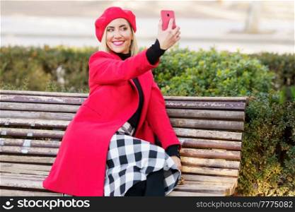 Delighted female in stylish red outfit taking self portrait on smartphone while sitting on wooden bench in park on autumn day. Cheerful woman taking selfie in park