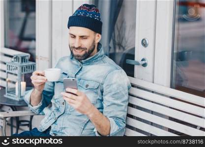 Delighted happy male with thick beard and mustache, reads positive news on cell phone, enjoys recreation time, drinks tea in coffee shop, has happy expression. Smiling handsome man uses cellular