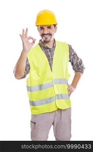 Delighted male builder in yellow helmet and reflective vest standing with hand on waist on white background and showing okay sign while looking at camera. Content male contractor showing OK gesture