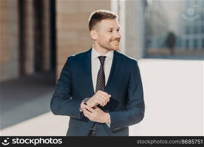 Delighted male CEO wears elegant formal suit, stands outdoor on street, has gentle positive smile, rejoices success in his career. Successful young businessman finishes work on business project
