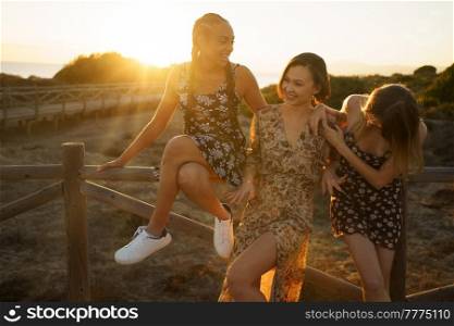Delighted multiracial female friends in sundresses standing near wooden barrier on waterfront on sunny summer day in coastal area against cloudless sky. Happy diverse girlfriends near wooden fence