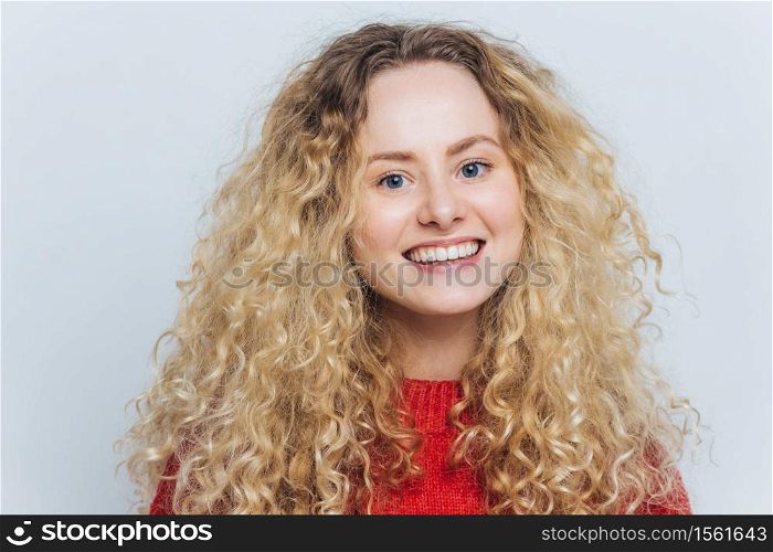 Delighted pleased adorable woman with curly blonde hair and warm blue eyes, being in good mood as recieves compliment, isolated over white background. Glad happy female smiles joyfully at camera
