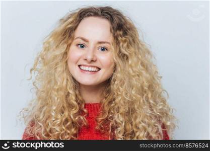 Delighted pleased adorable woman with curly blonde hair and warm blue eyes, being in good mood as recieves compliment, isolated over white background. Glad happy female smiles joyfully at camera