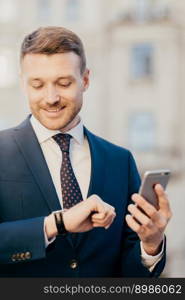 Delighted unshaven male corporation owner looks happily at wristwatch, checks time, recieves incoming sms message on smart phone, updates profile of bank account, uses free interent connection