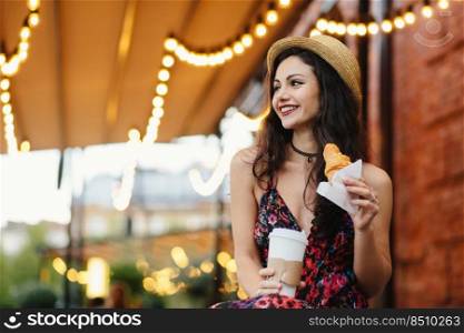 Delightful brunette female with pleasant appearance wearing summer hat and dress holding croissant and takeaway coffee, resting at terrace looking aside with happy expression. People, recreation