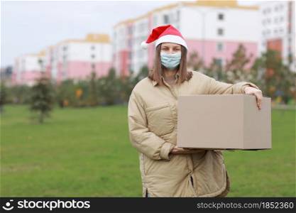 Delivery girl in a red Santa Claus hat and medical protective mask holds a big box outdoor. New Year&rsquo;s Christmas parcel in box, delivery online store in Quarantine time. Service coronavirus. Delivery girl in a red Santa Claus hat and medical protective mask holds a big box outdoor. New Year&rsquo;s Christmas parcel in box, delivery online store in Quarantine time. Service coronavirus.