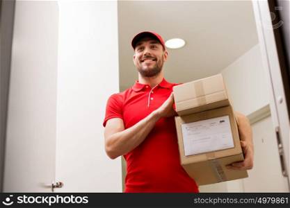 delivery, mail, people and shipment concept - happy man in red uniform with parcel boxes in corridor at open customer door. delivery man with parcel boxes at customer door
