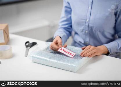 delivery, mail service, and shipment concept - close up of woman sticking fragile mark to parcel in protective bubble wrap at post office. woman sticking fragile mark to wrap at post office