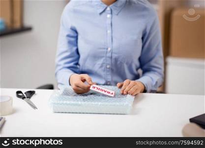 delivery, mail service, and shipment concept - close up of woman sticking fragile mark to parcel in protective bubble wrap at post office. woman sticking fragile mark to wrap at post office
