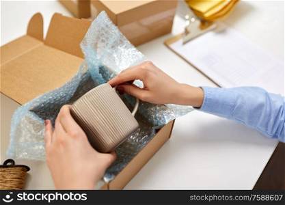 delivery, mail service, people and shipment concept - close up of female hands packing mug into parcel box and it wrapping into protective bubble wrap at post office. hands packing mug to parcel box at post office
