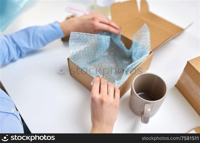delivery, mail service, people and shipment concept - close up of female hands packing mug into parcel box and it wrapping into protective bubble wrap at post office. hands packing mug to parcel box at post office