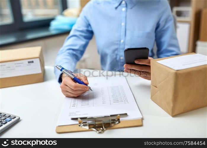 delivery, mail service, people and shipment concept - close up of woman with smartphone and parcel boxes filling checklist on clipboard at post office. woman with smartphone and clipboard at post office